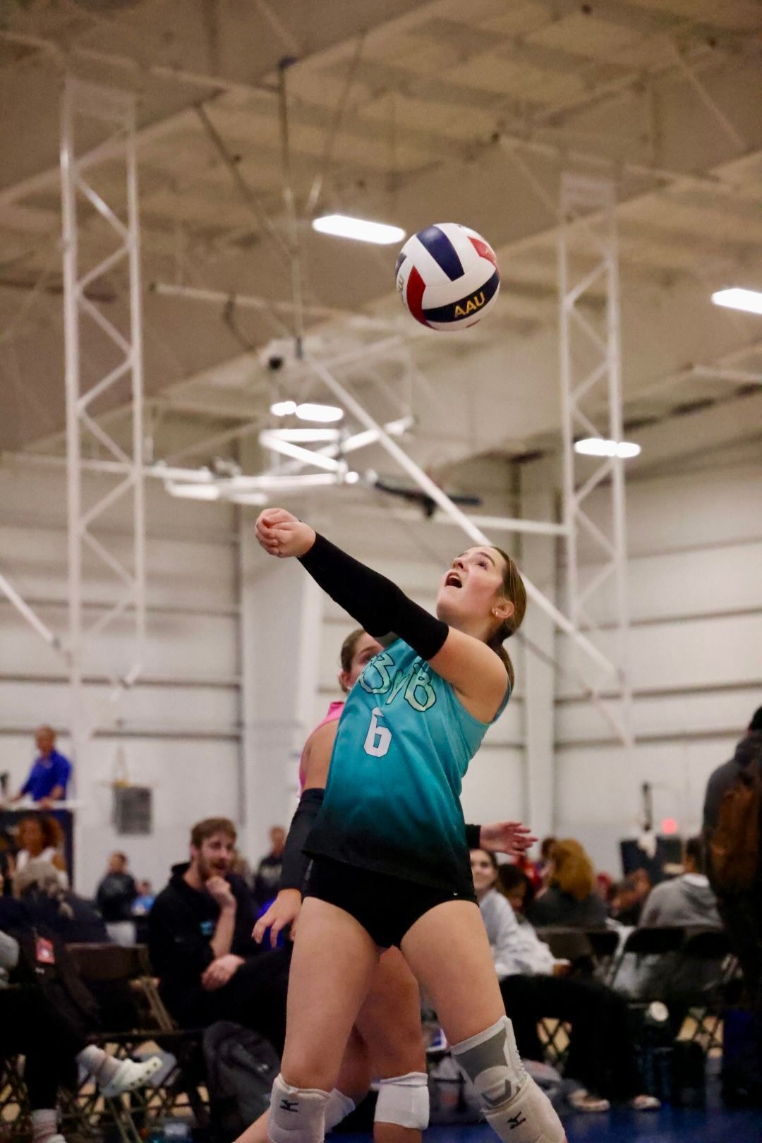 Volleyball player in teal uniform sets the ball indoors, eyes up, mouth open.