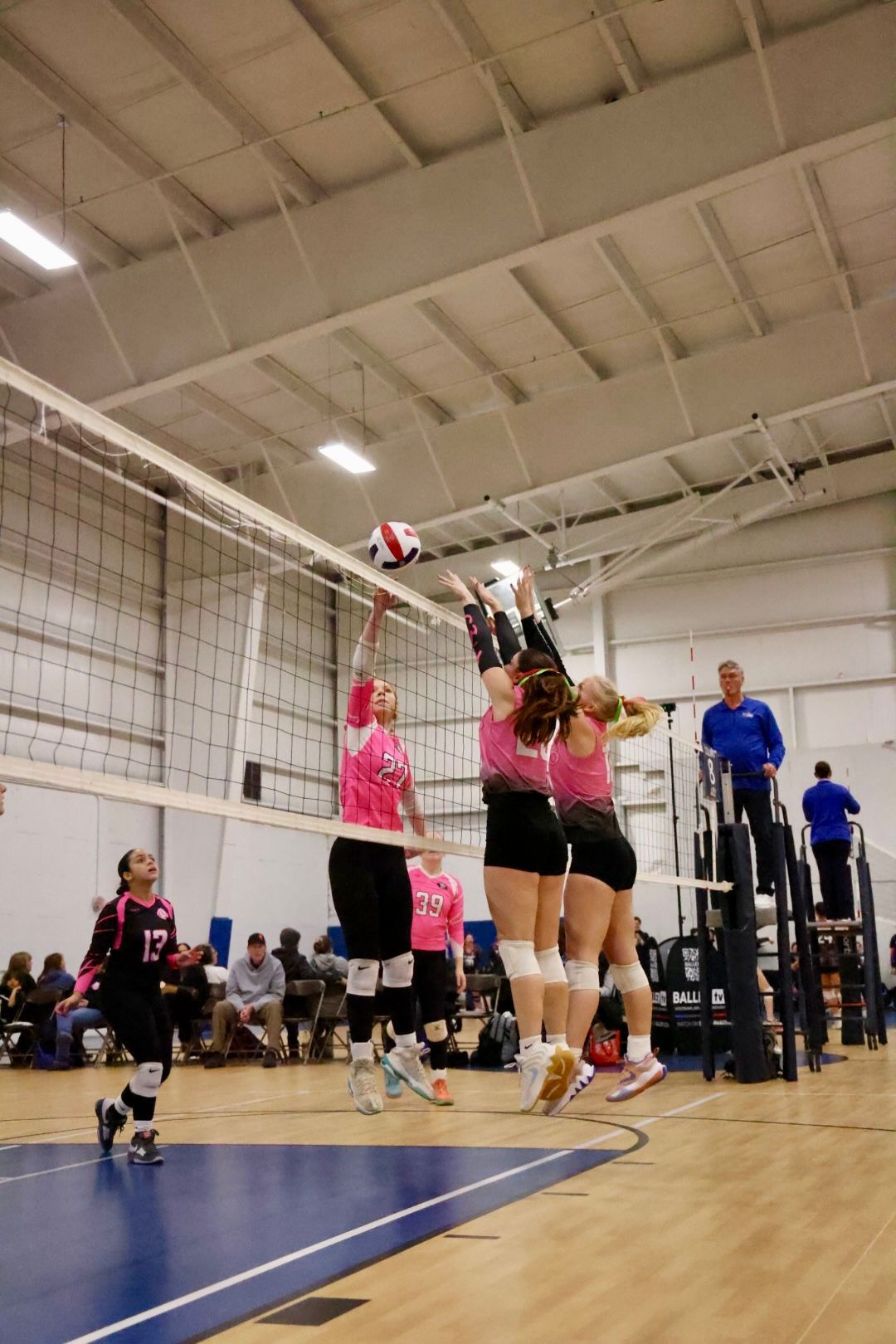 Volleyball players in pink and black uniforms blocking a spike at a net in a gymnasium.