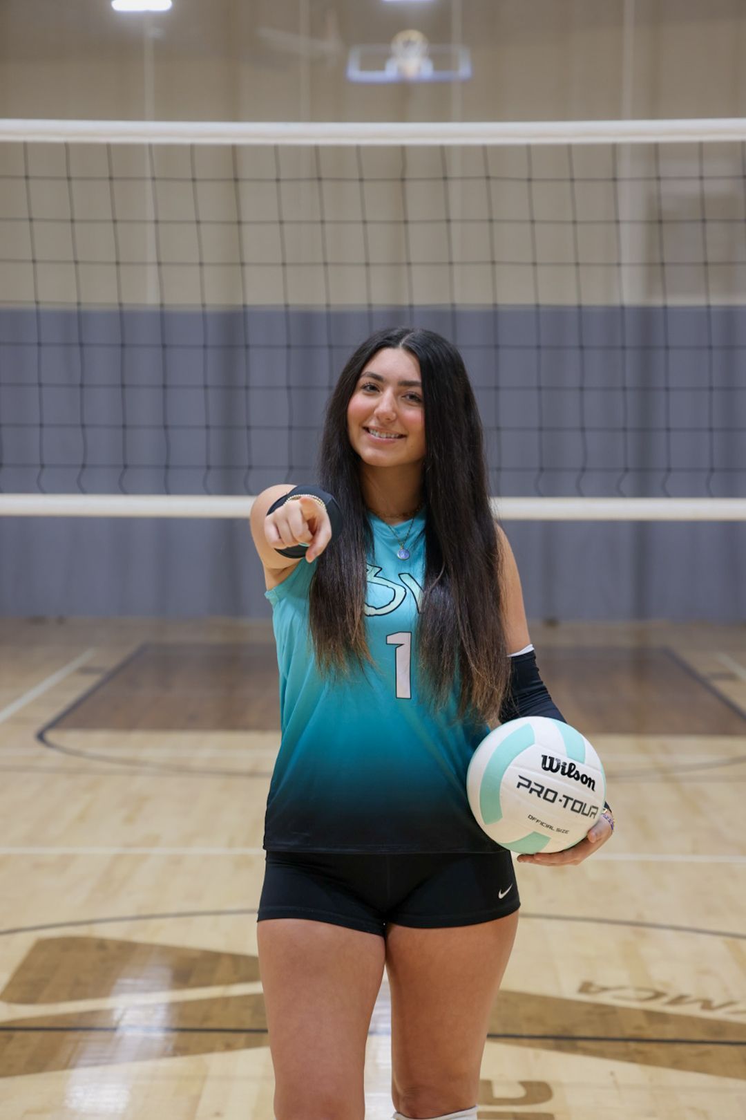 A person in a turquoise and black volleyball jersey holds a ball, pointing forward on a court in front of a net.