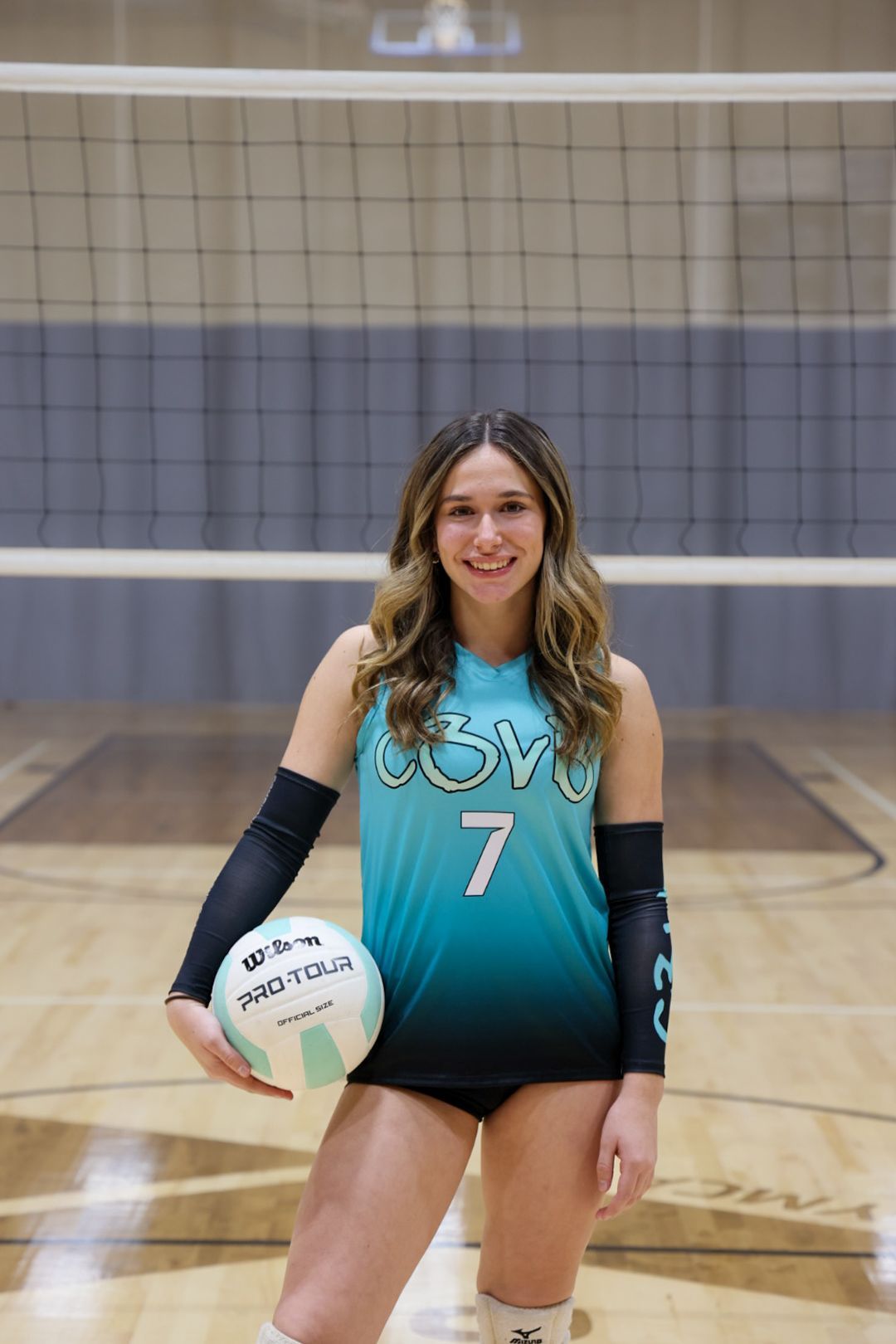 Volleyball player wearing a teal and black jersey with number 7 stands on a court holding a volleyball.