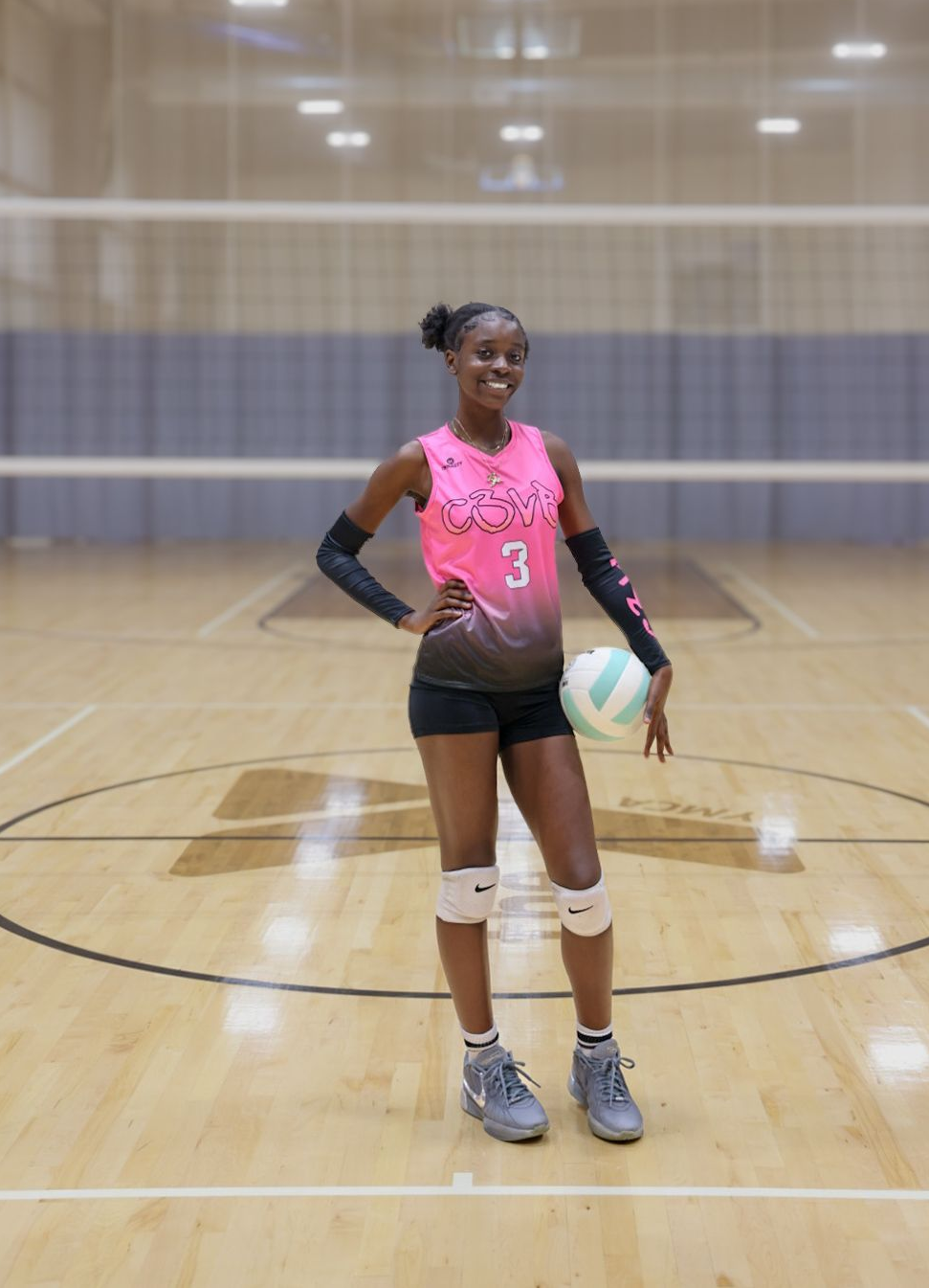 A volleyball player in a pink and black uniform stands on a court, holding a volleyball and smiling.