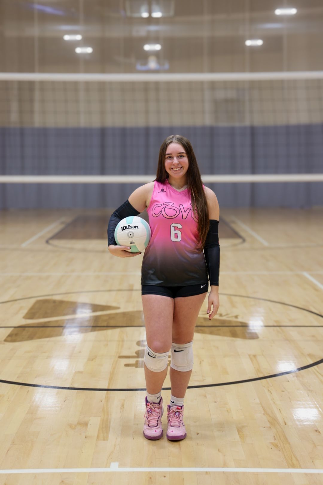 A smiling volleyball player in a pink and black jersey holds a ball, standing on an indoor court with nets in the back.
