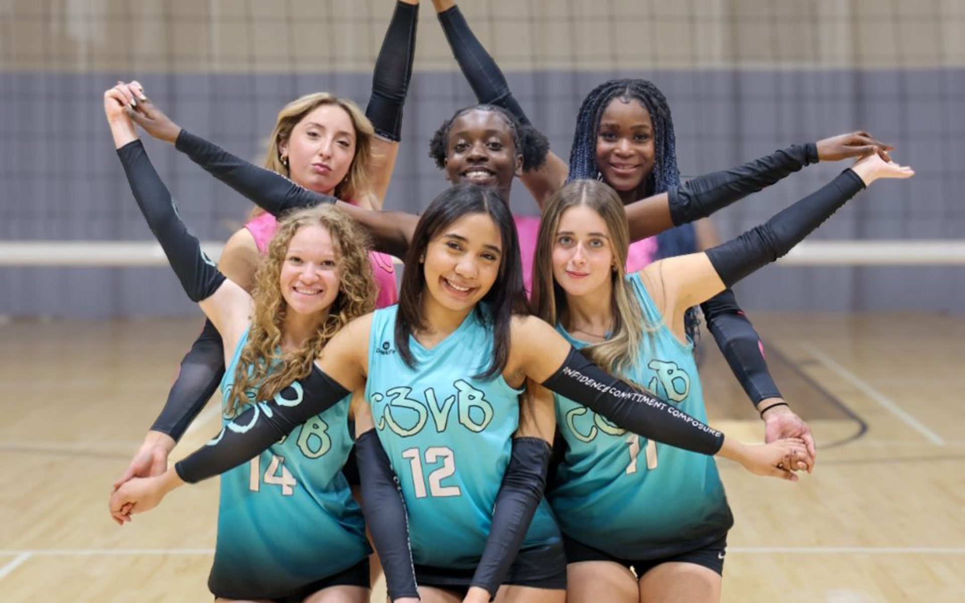 Volleyball team in teal and pink jerseys forms a star shape with their arms. Gym setting.