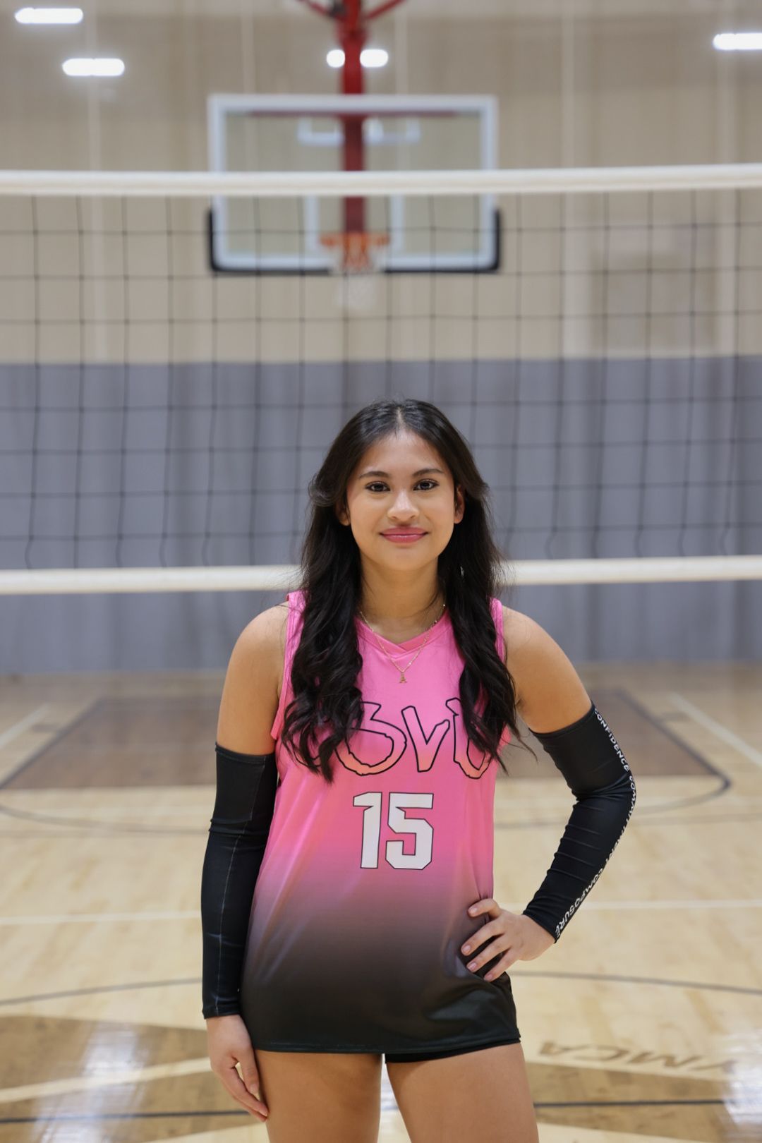 A person in a pink and black jersey with the number 15 stands on a volleyball court in front of a net and basketball hoop.