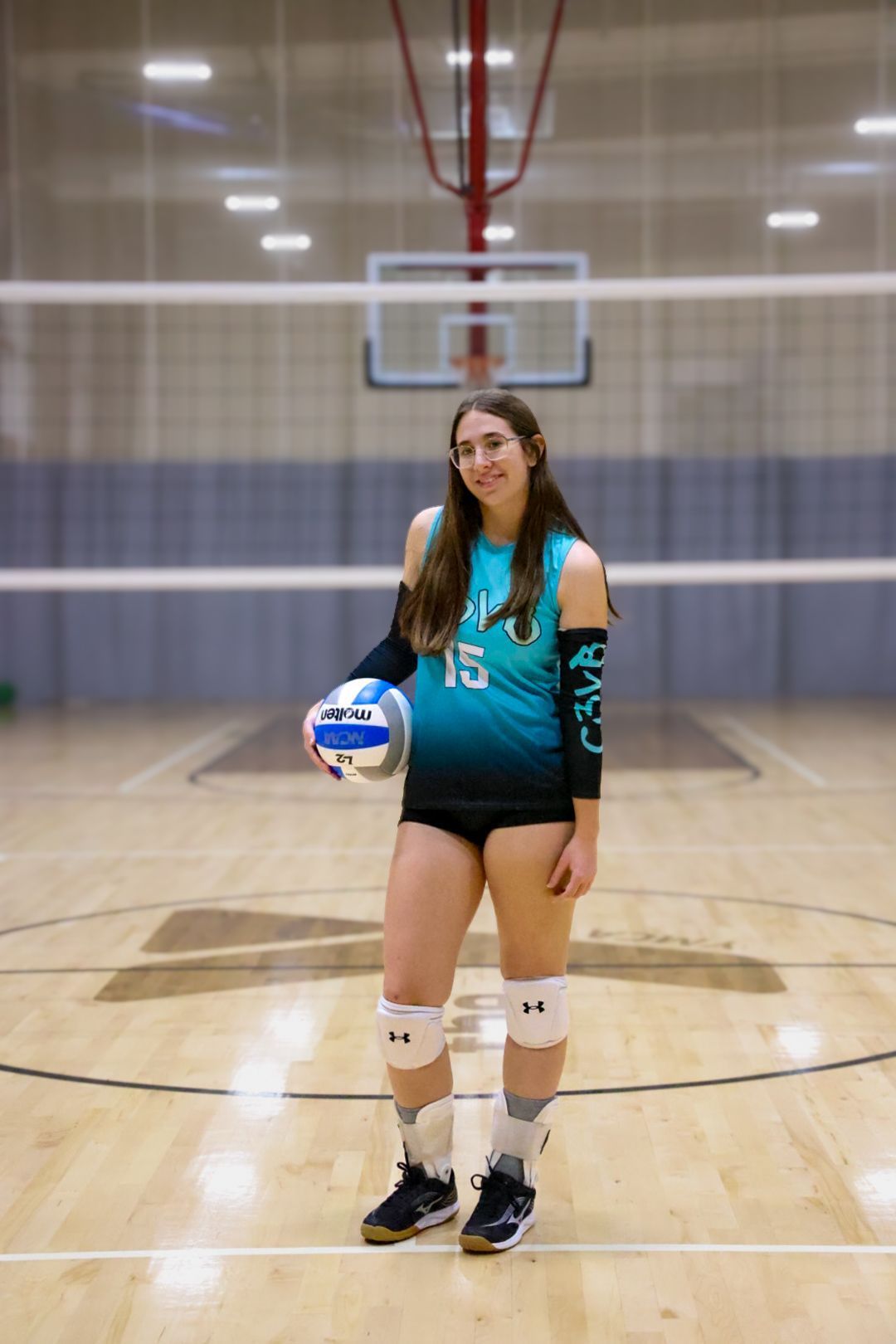 A volleyball player stands on a court in a teal and black jersey holding a ball, wearing knee pads and an ankle brace.
