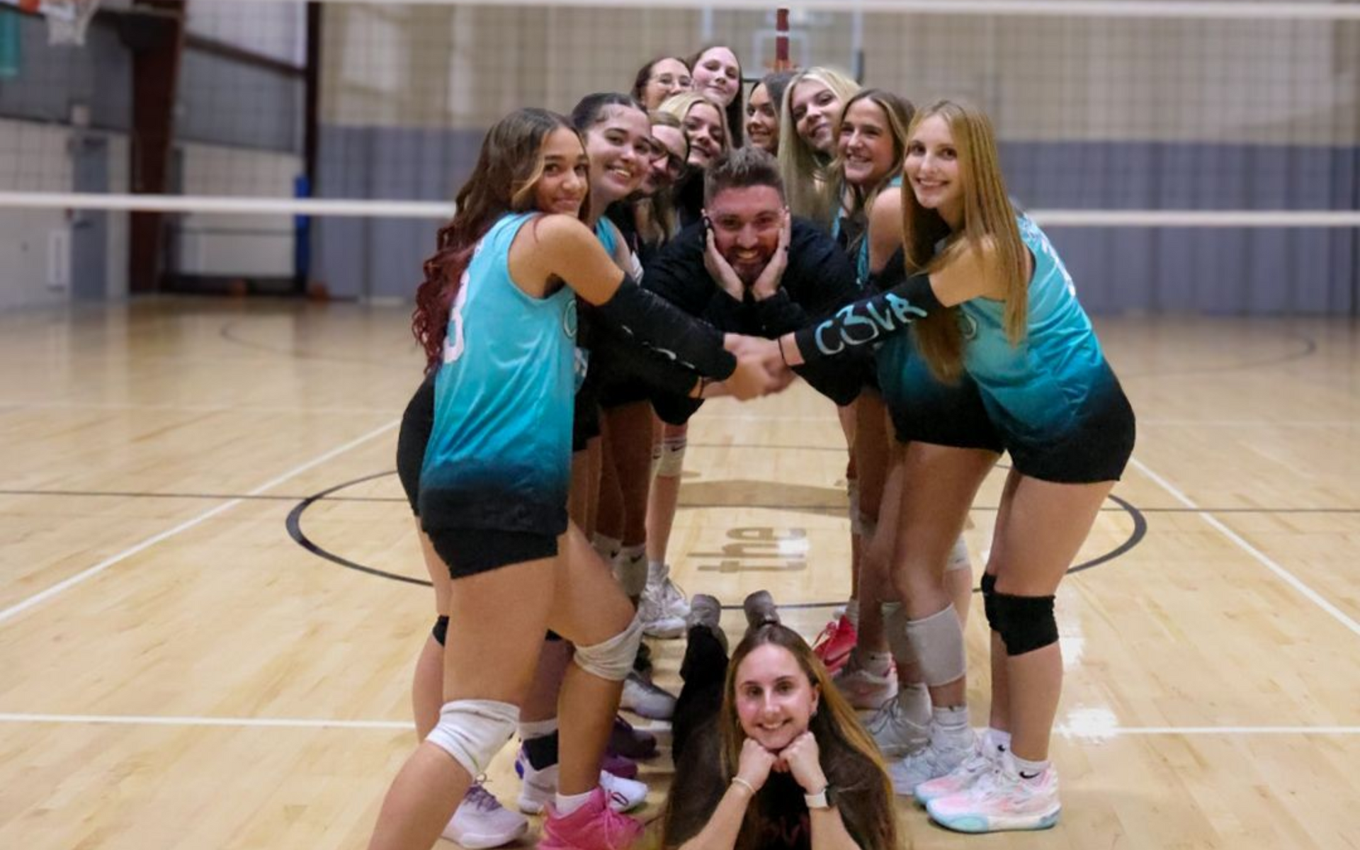 Volleyball team huddles together on court, hands in center, smiling.