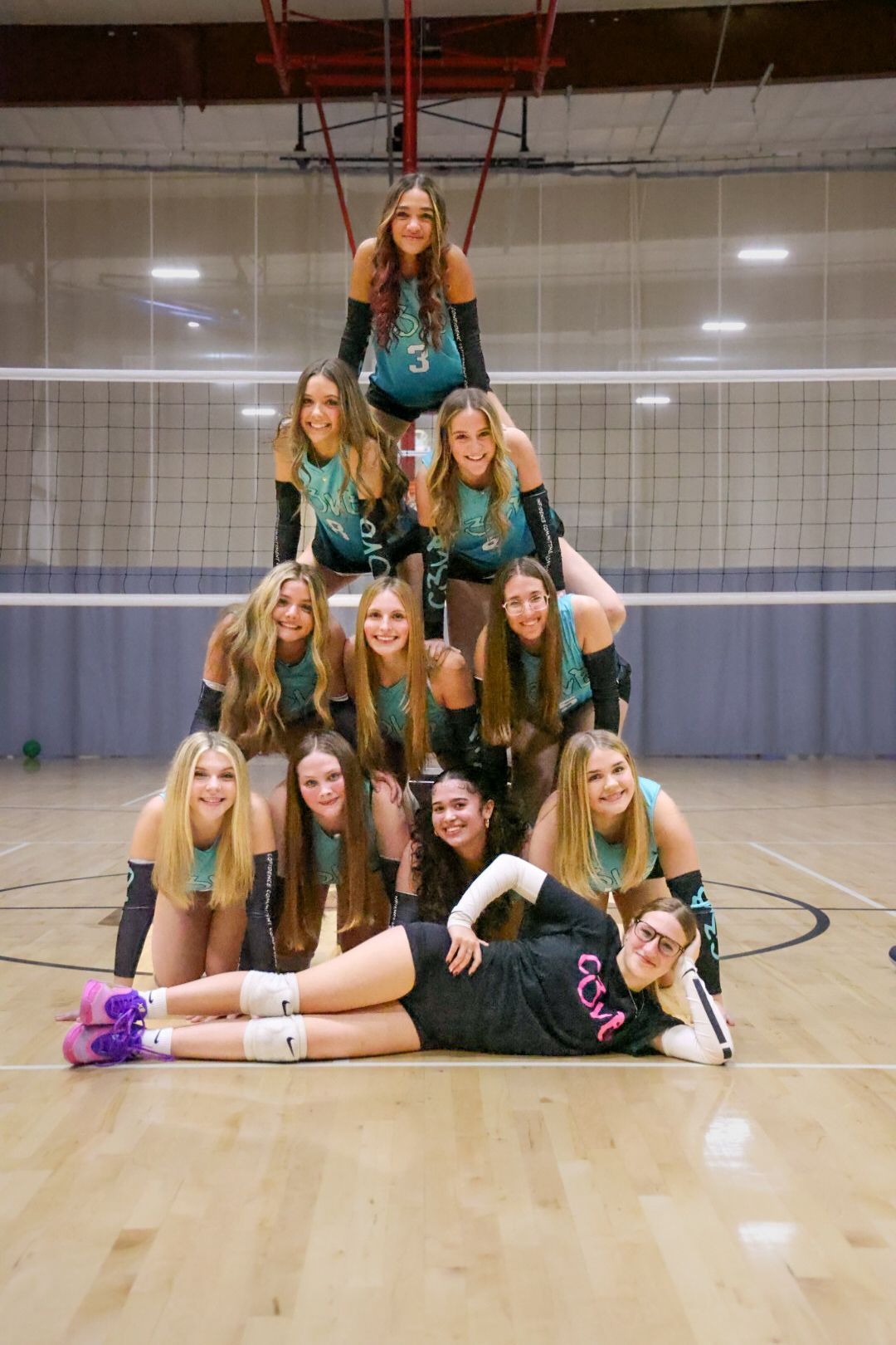 Volleyball team in matching teal uniforms, posing in pyramid formation on court.