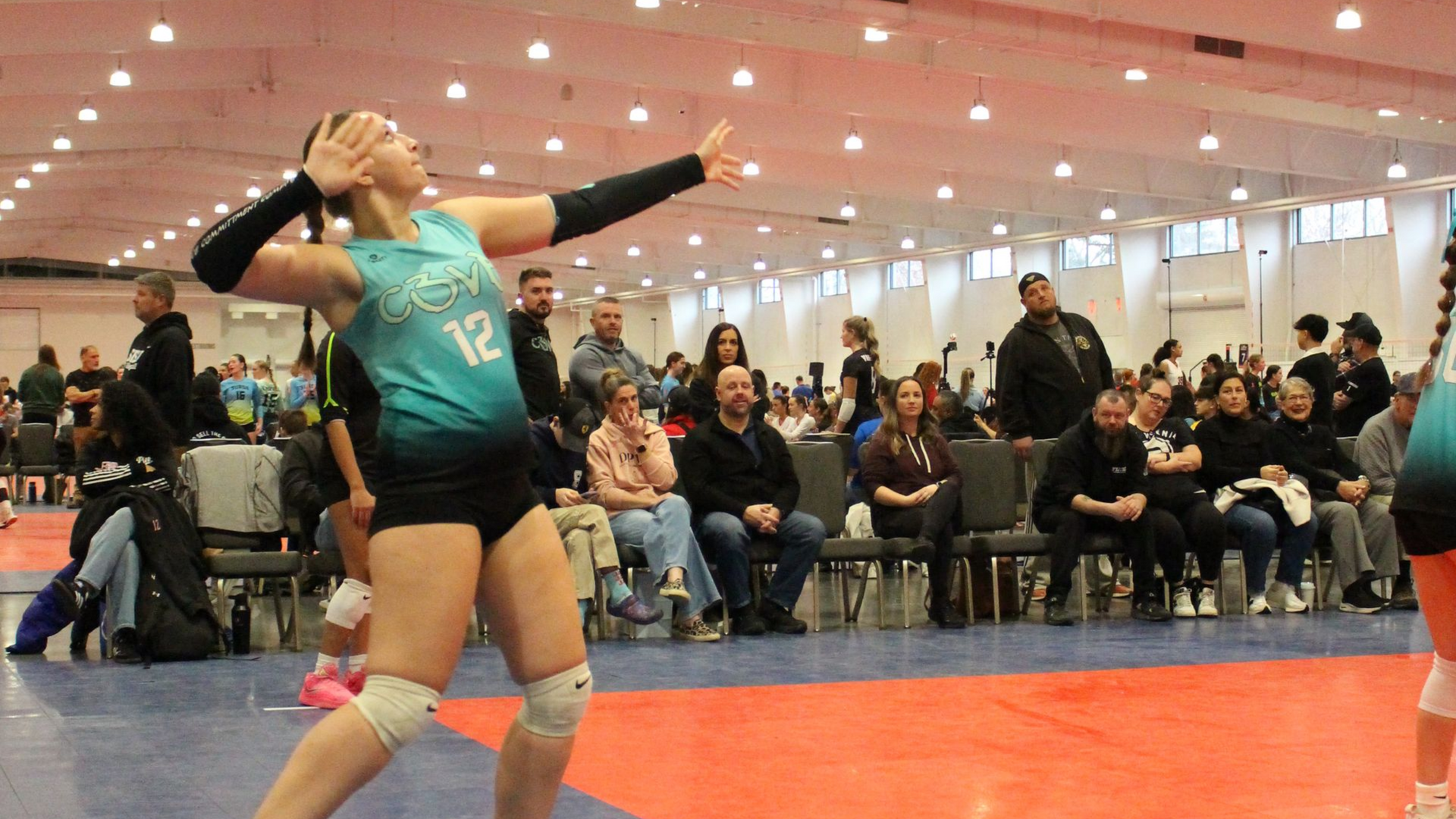 Volleyball player in teal jersey about to serve the ball in a large indoor venue, with spectators in the background.