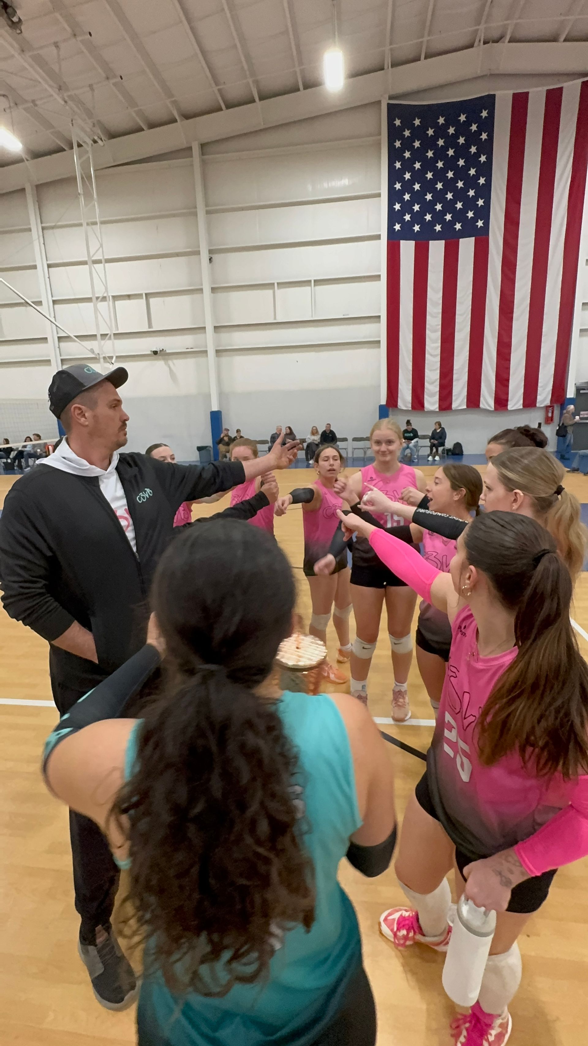 A coach gestures to a huddle of volleyball players on a court in front of a large American flag.