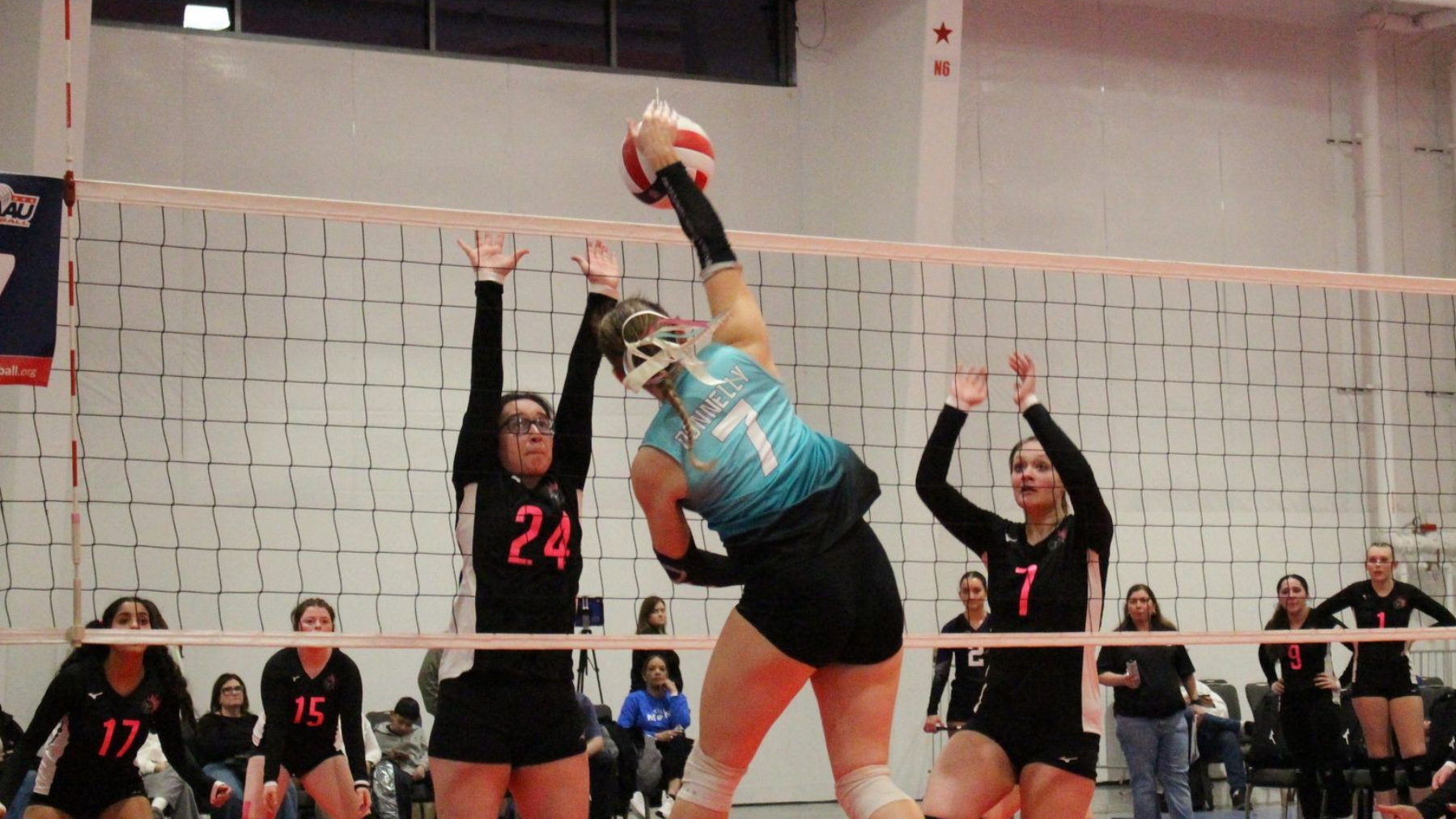 Volleyball player spikes the ball over the net, blocked by two opponents. Indoor court.