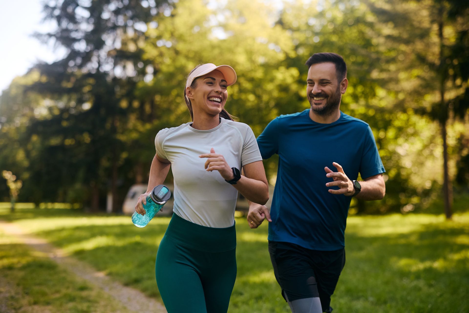 Two runners jogging on a sunny park trail, smiling and carrying water bottles