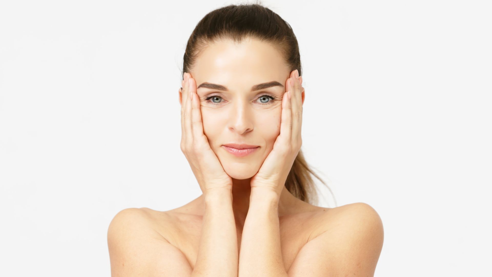Woman with fair skin and light eyes, touching her face, smiling slightly, neutral background.