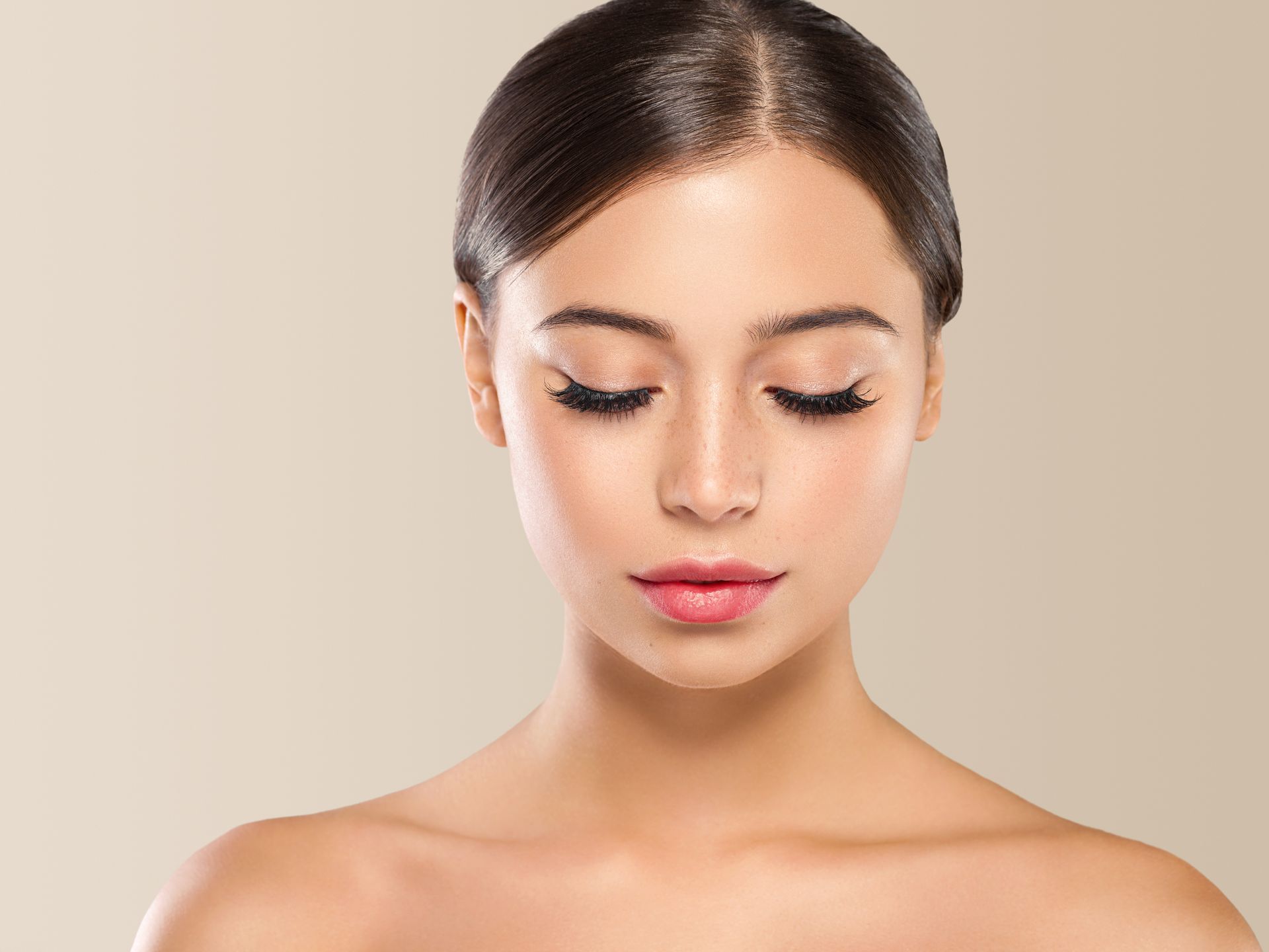 Woman with closed eyes, dark hair pulled back, wearing subtle makeup, beige background.