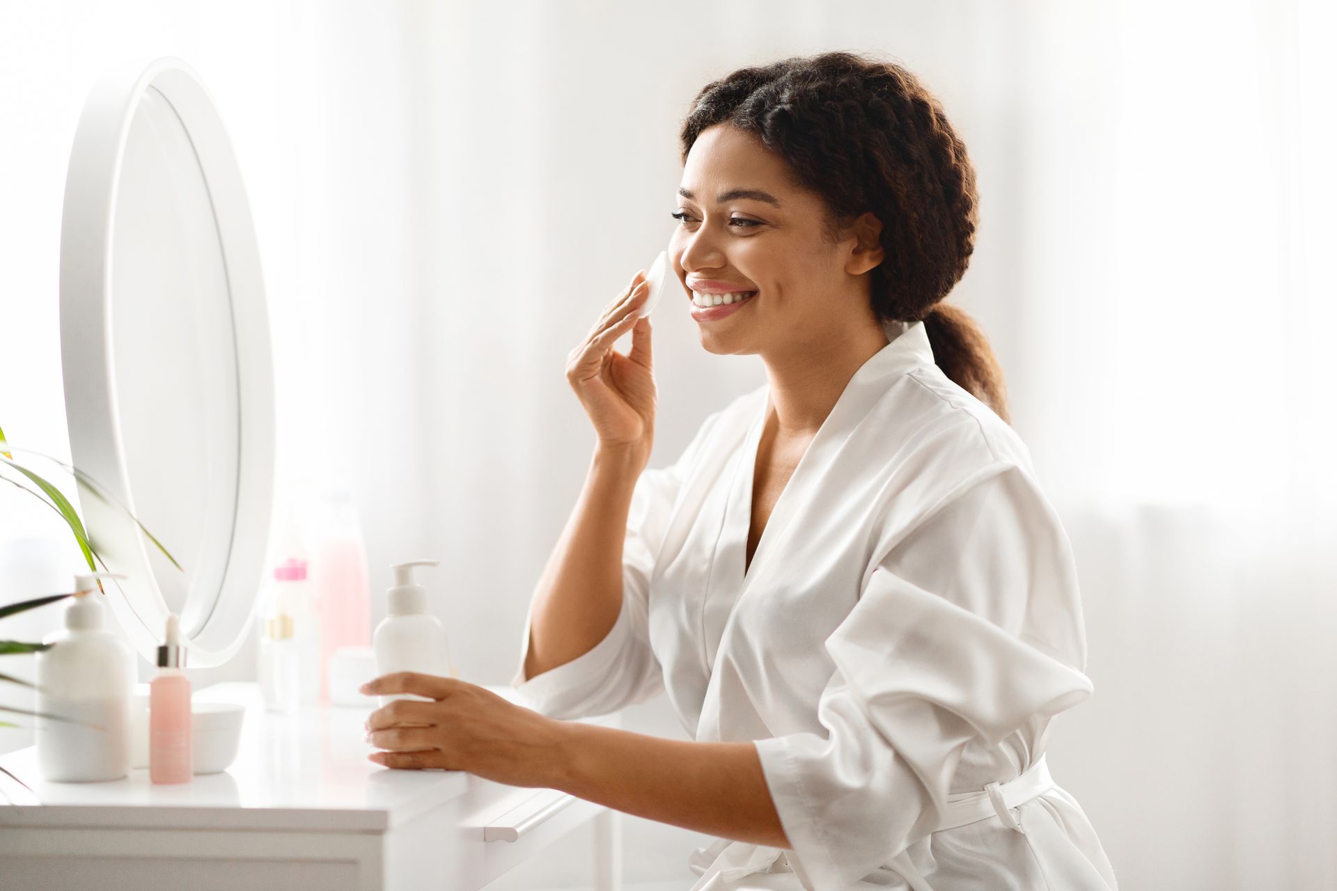 Woman in a white robe cleanses her face with a cotton pad in front of a mirror.