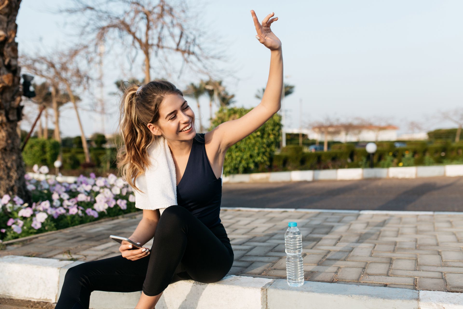 Woman in workout clothes waving, smiling outdoors with a phone, towel, and water bottle.