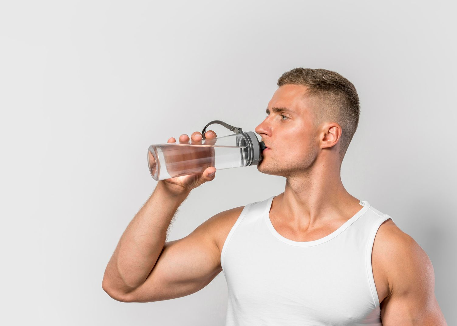 Man in white tank top drinks from a clear water bottle, against a white background.