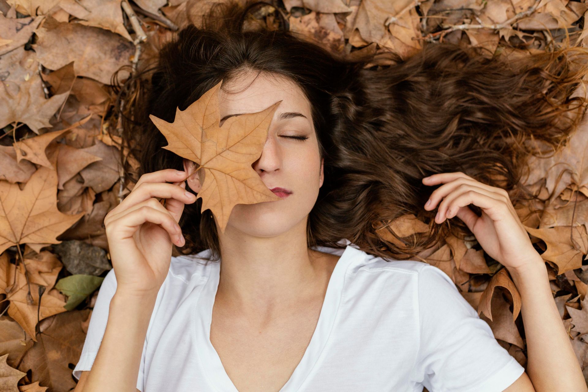 Woman lying in autumn leaves, covering her eye with a leaf; wearing a white shirt, brown hair.