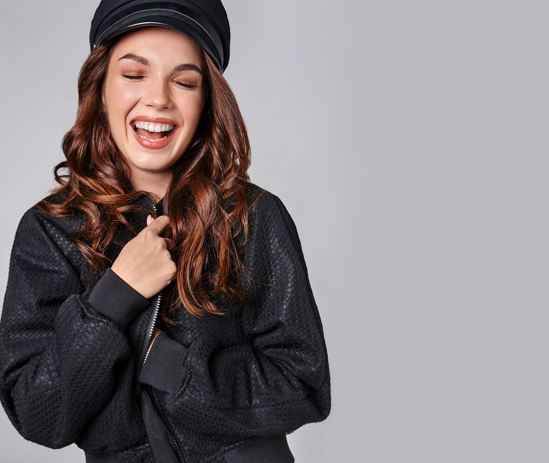 Woman laughing, wearing a black cap and jacket, against a gray background.