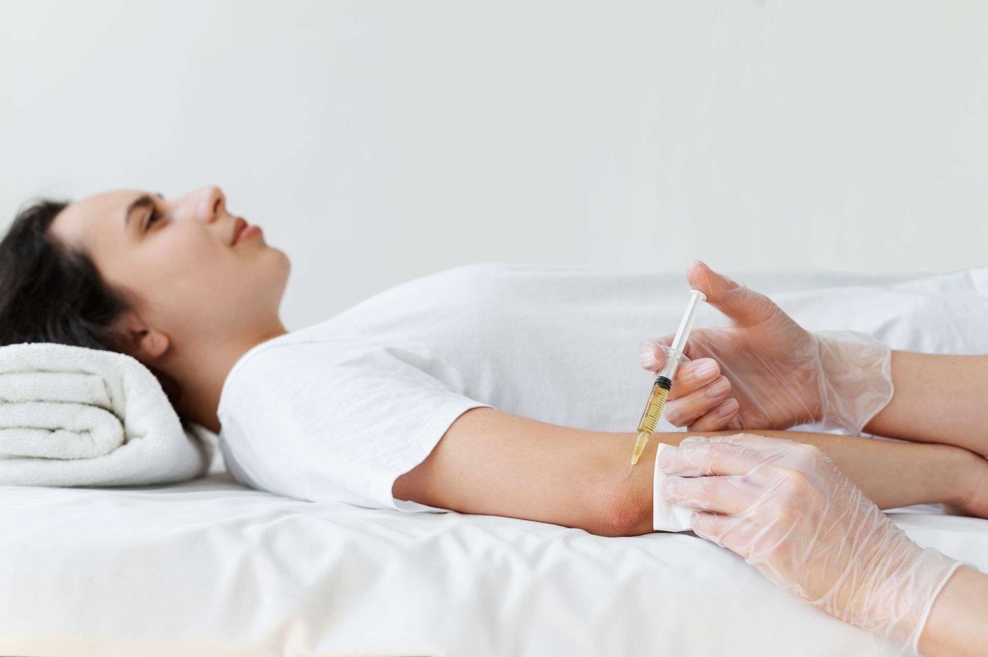 Woman receiving an elbow injection in a white-walled room, with a healthcare professional administering the treatment.