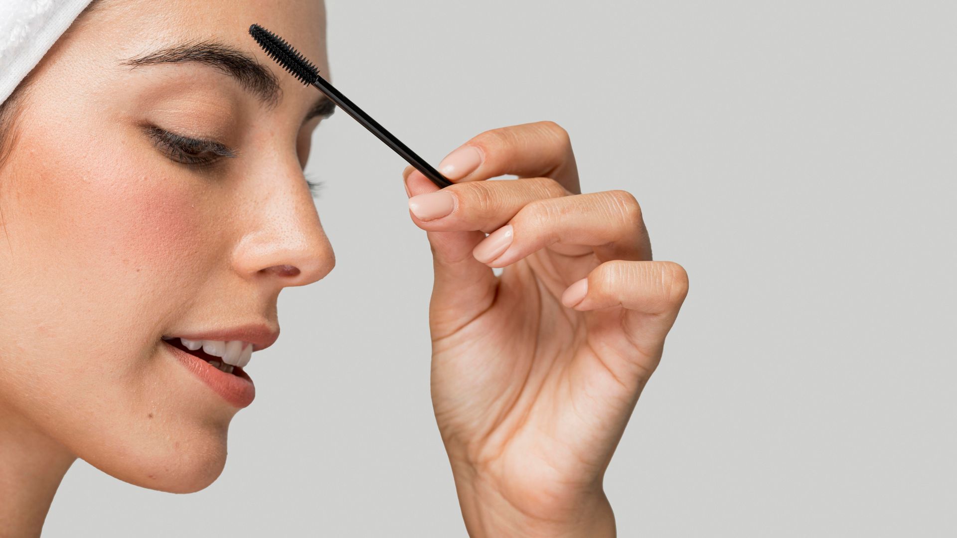 Woman brushes her eyebrow with a brow brush. She is smiling, with a white head wrap.
