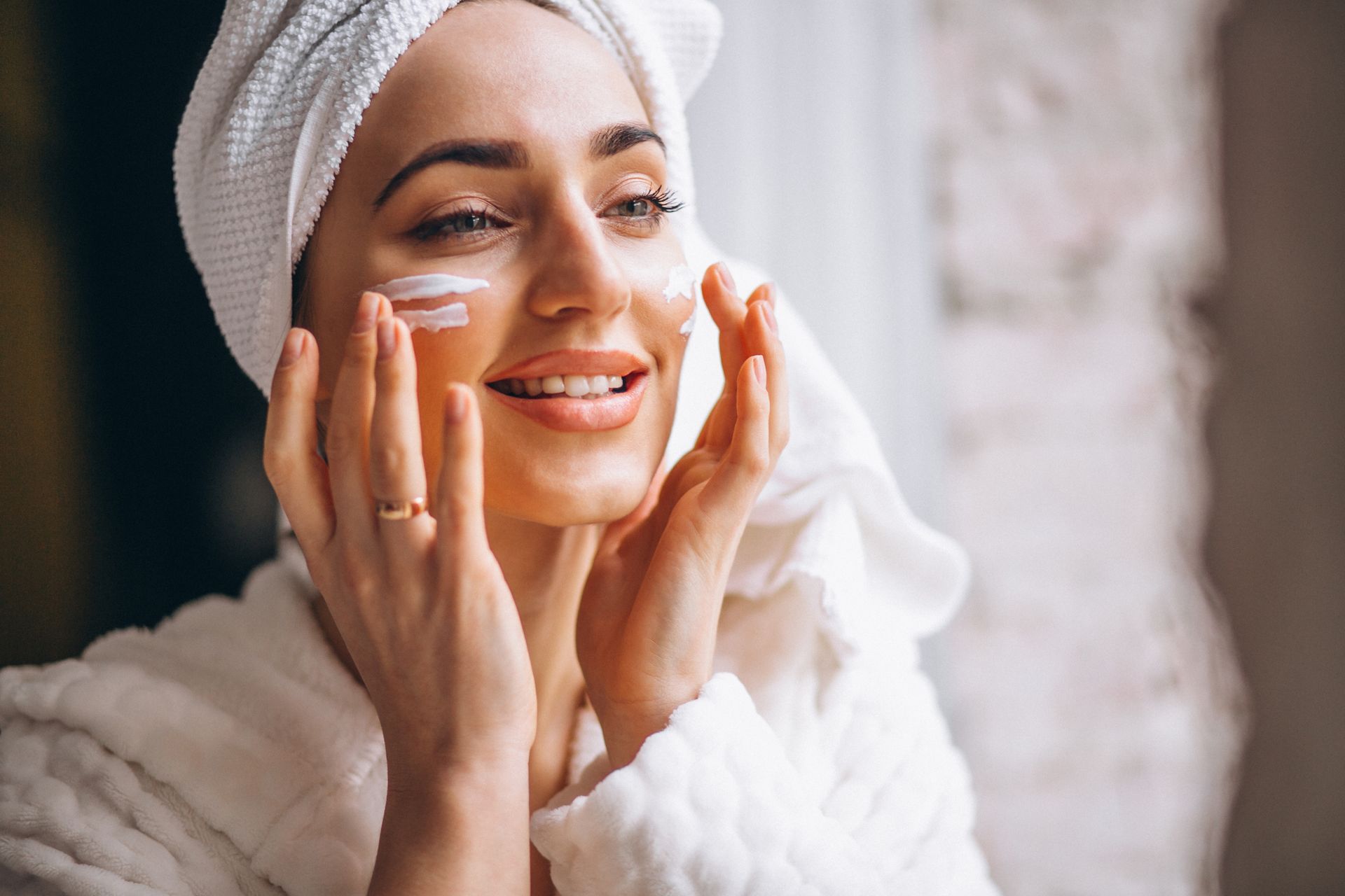 Woman in bathrobe and towel applying cream to her face, smiling, near a window.