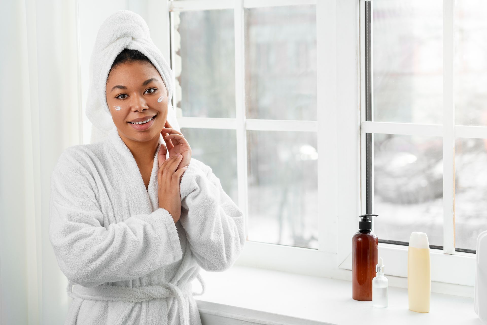 Woman in bathrobe and towel, smiling, near window with skincare products.