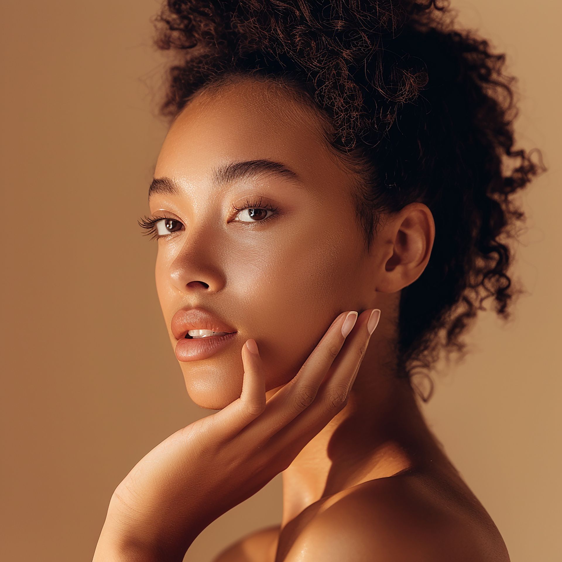 Woman with radiant skin, touching her face, looking towards the camera with a soft smile. Beige background.