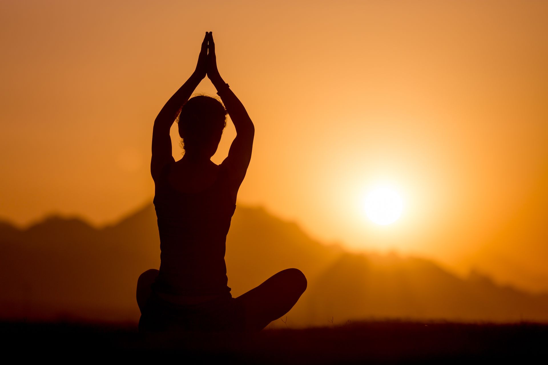 Silhouette of a person in yoga pose with hands raised, against a sunset with mountains.