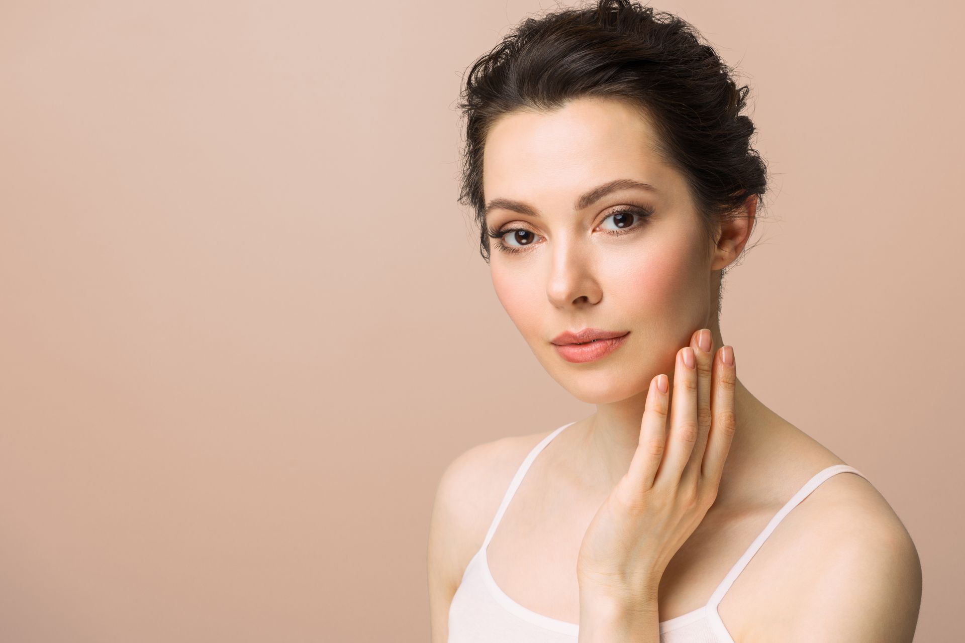 Woman in white tank top touches her cheek, looking at the viewer. Brown hair updo, neutral background.