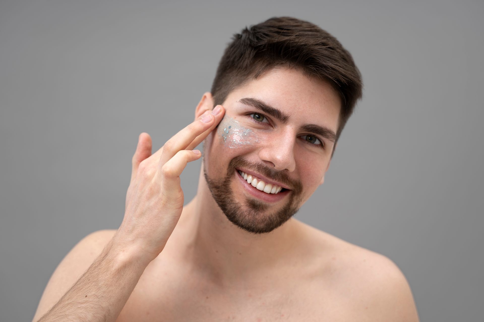 Smiling man applying skincare product to his face, against a gray background.