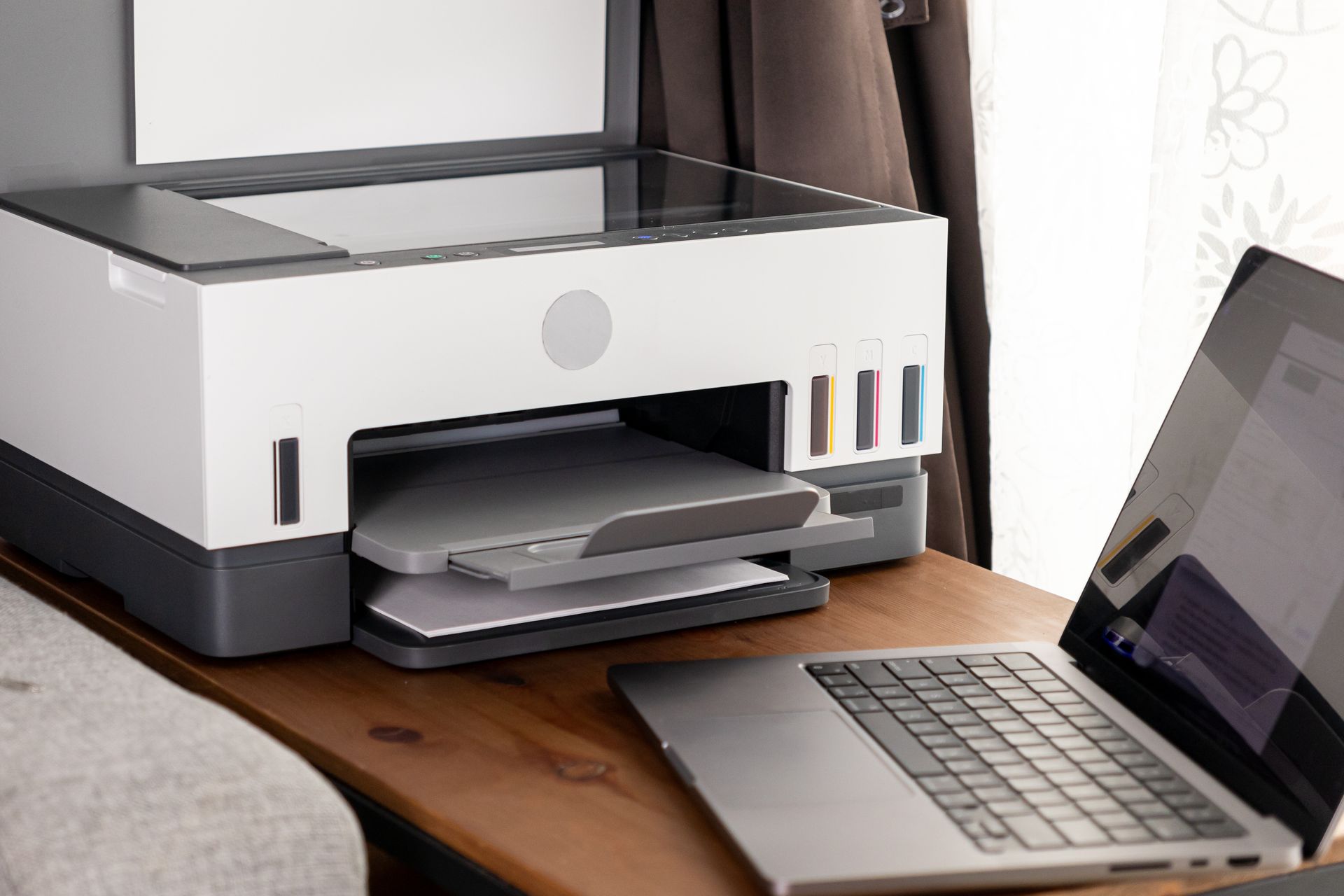 A white and gray inkjet printer sits on a wooden desk next to an open laptop.
