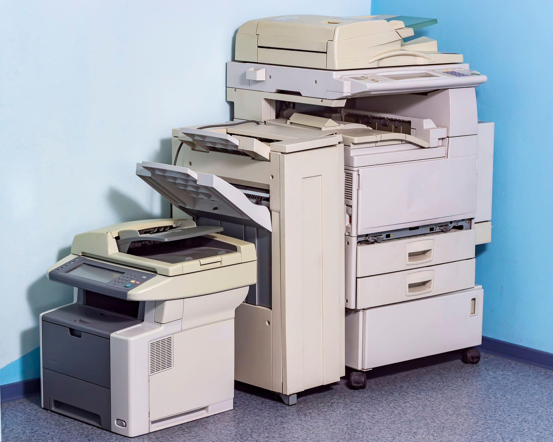 Three office copy machines of varying sizes, colored beige and grey, placed in the corner of a room with light blue walls.