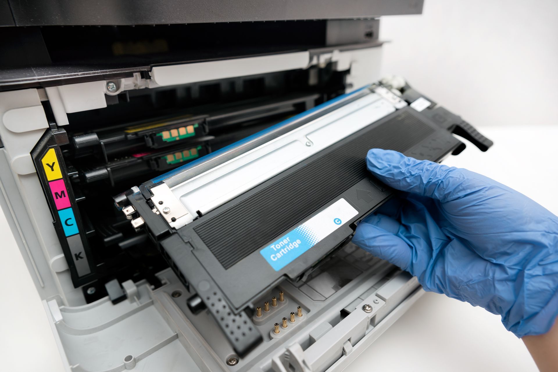 A gloved hand inserting a blue toner cartridge into a color laser printer.