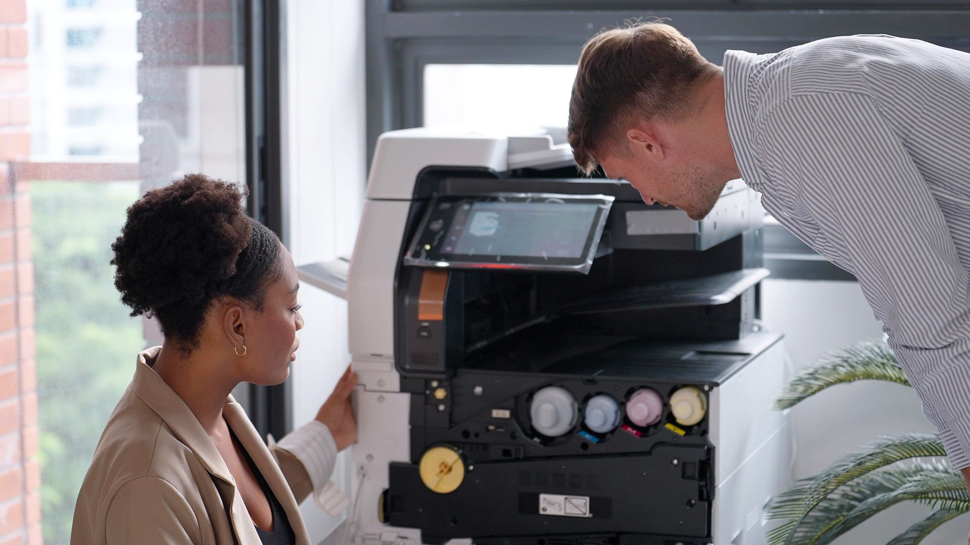 Two office workers examine the inside of a large printer in a well-lit office with a window.