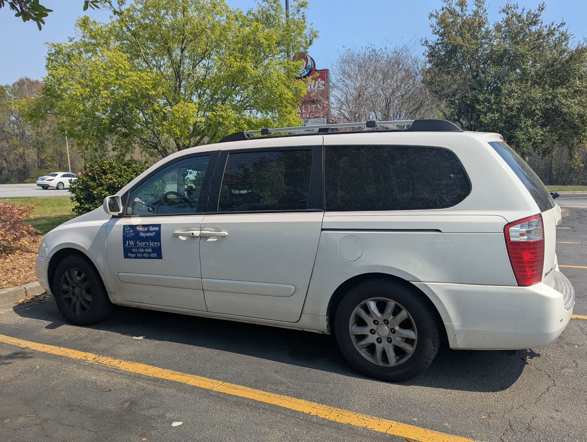 A white minivan with a roof rack and business signage on the driver-side door, parked in an outdoor parking lot.
