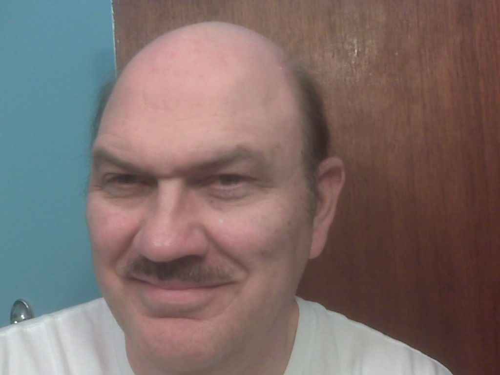 A person with a mustache smiling at the camera against a wooden door and a blue wall.