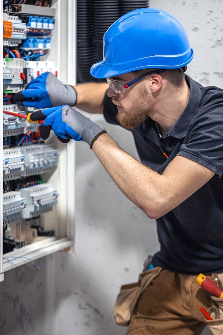 Electrician wearing a hard hat and gloves working on a circuit panel with tools.