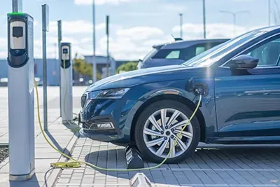Blue electric car charging at a station, with a charging cable connected. Sunny outdoor setting.