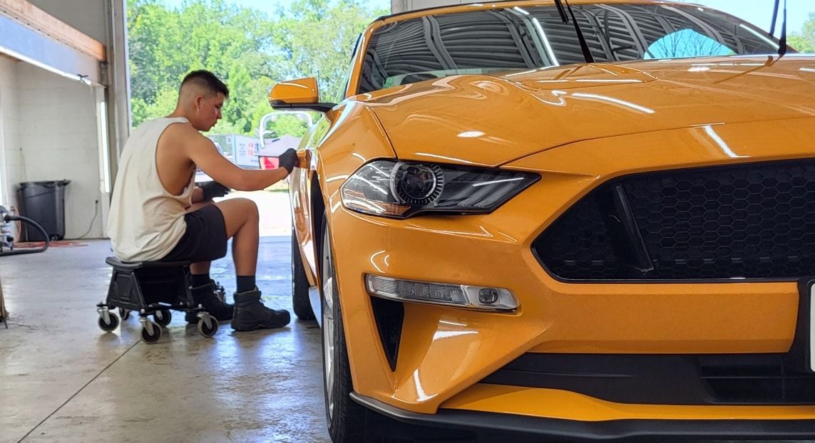 A man is sitting on a stool in front of a yellow mustang.