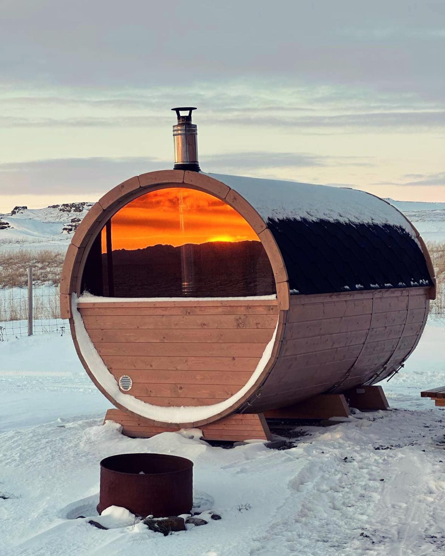 En tønnesauna i arktisk vinterlandskap, i vinduets refleksjon kan man se solnedgang.