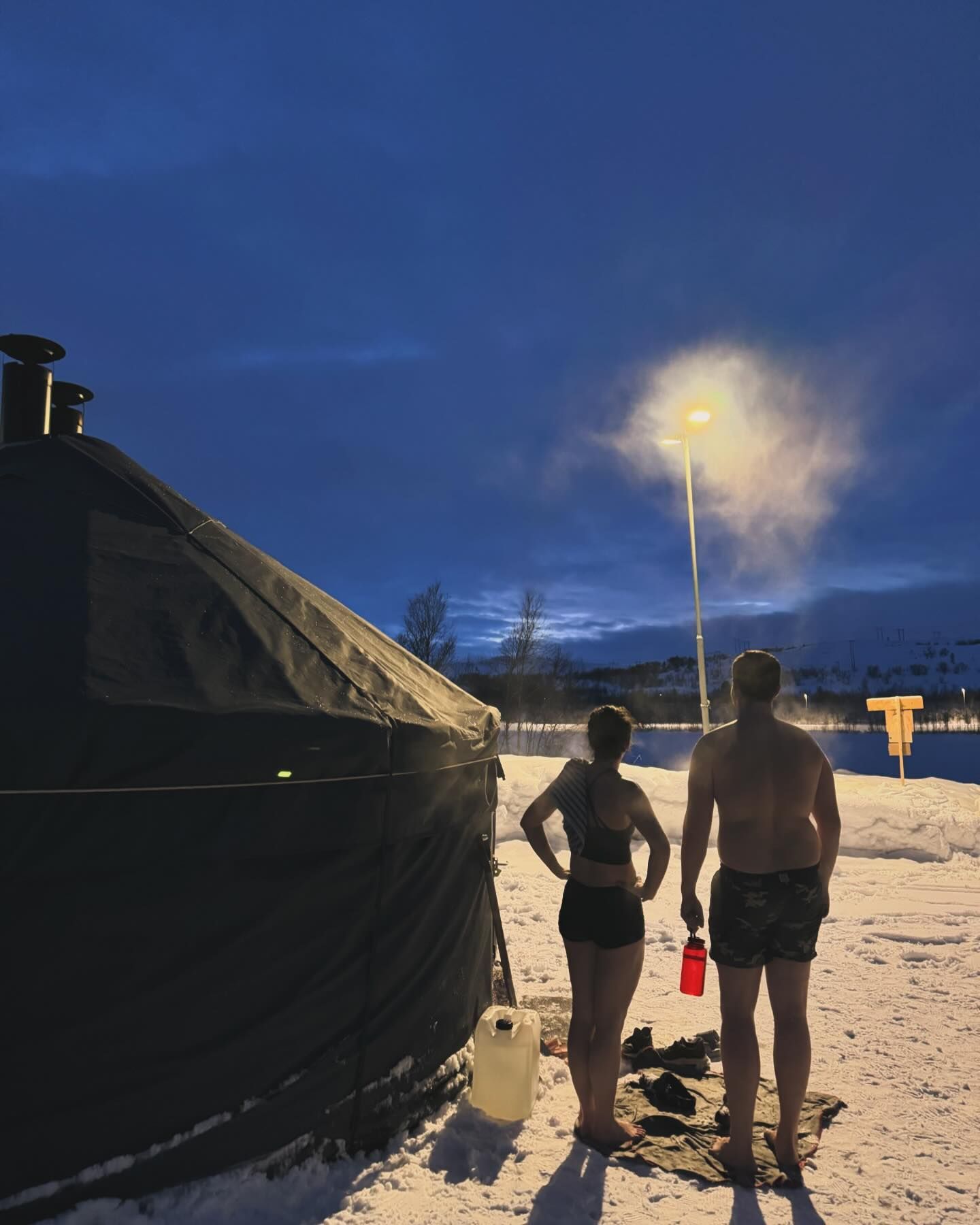 En kvinne og en mann står i badetøy utenfor en badstujurt på vinterstid.