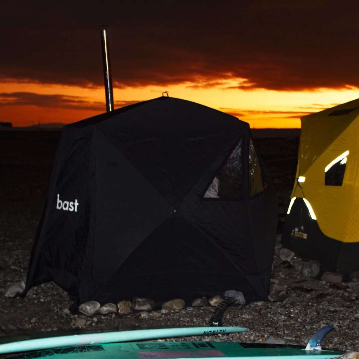 Teltsauna i solnedgang på en strand, det er et surfebrett i forgrunnen.