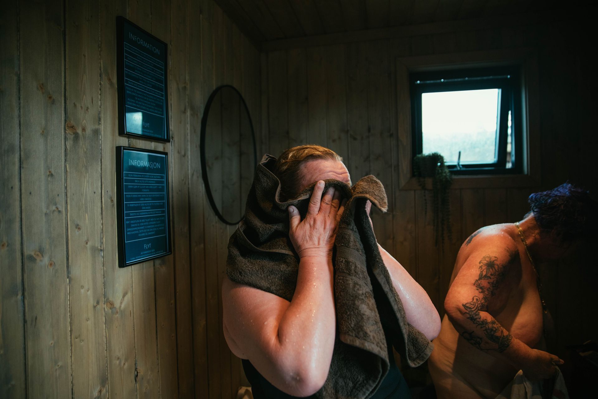 To kvinner med håndkle i et sauna-omkledningsrom – én tørker seg, én kler på seg.