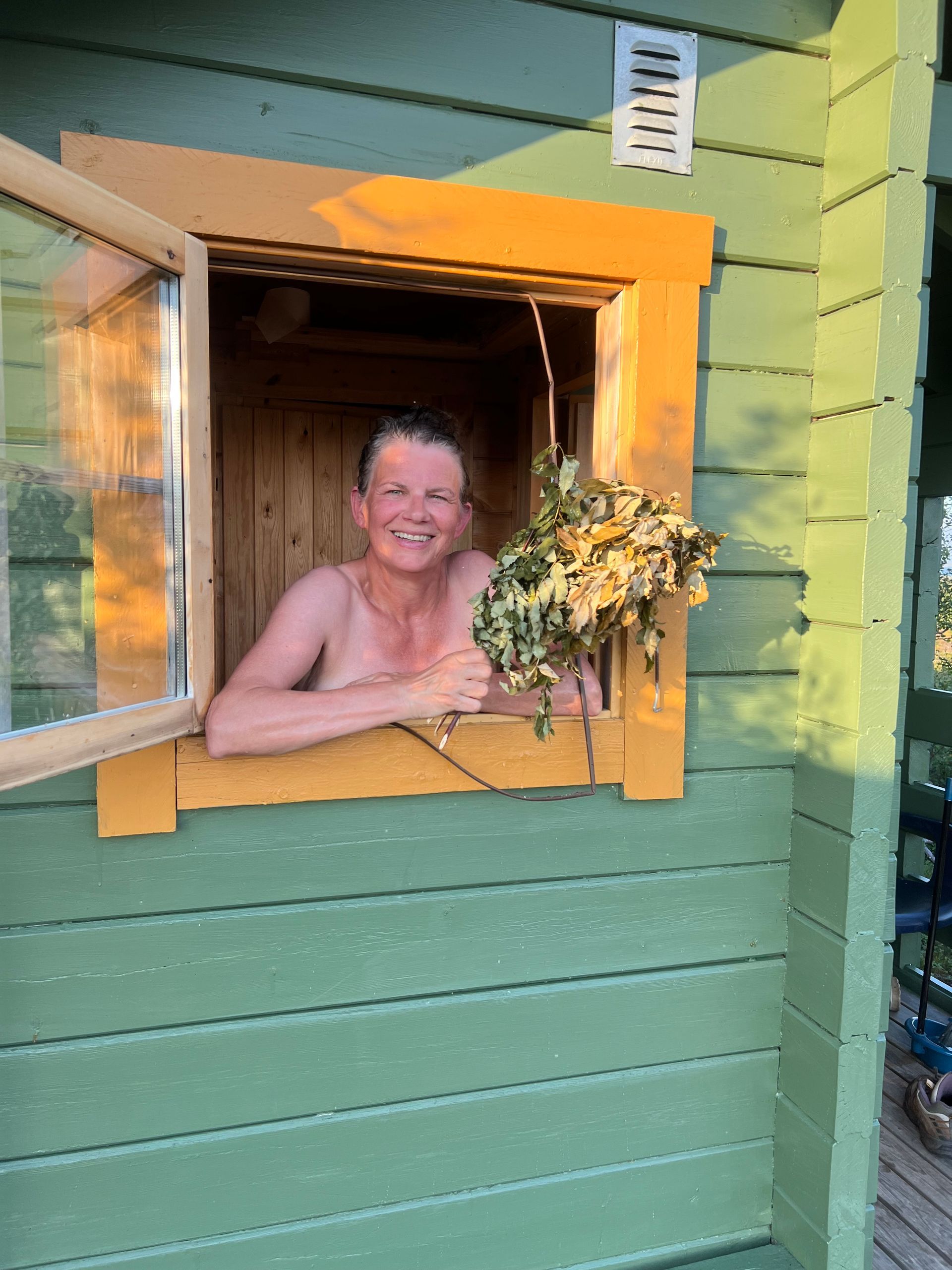 En kvinne titter ut av vinduet på en sauna i solnedgangen, hun smiler bredt og holder en badstulime av bjørk.