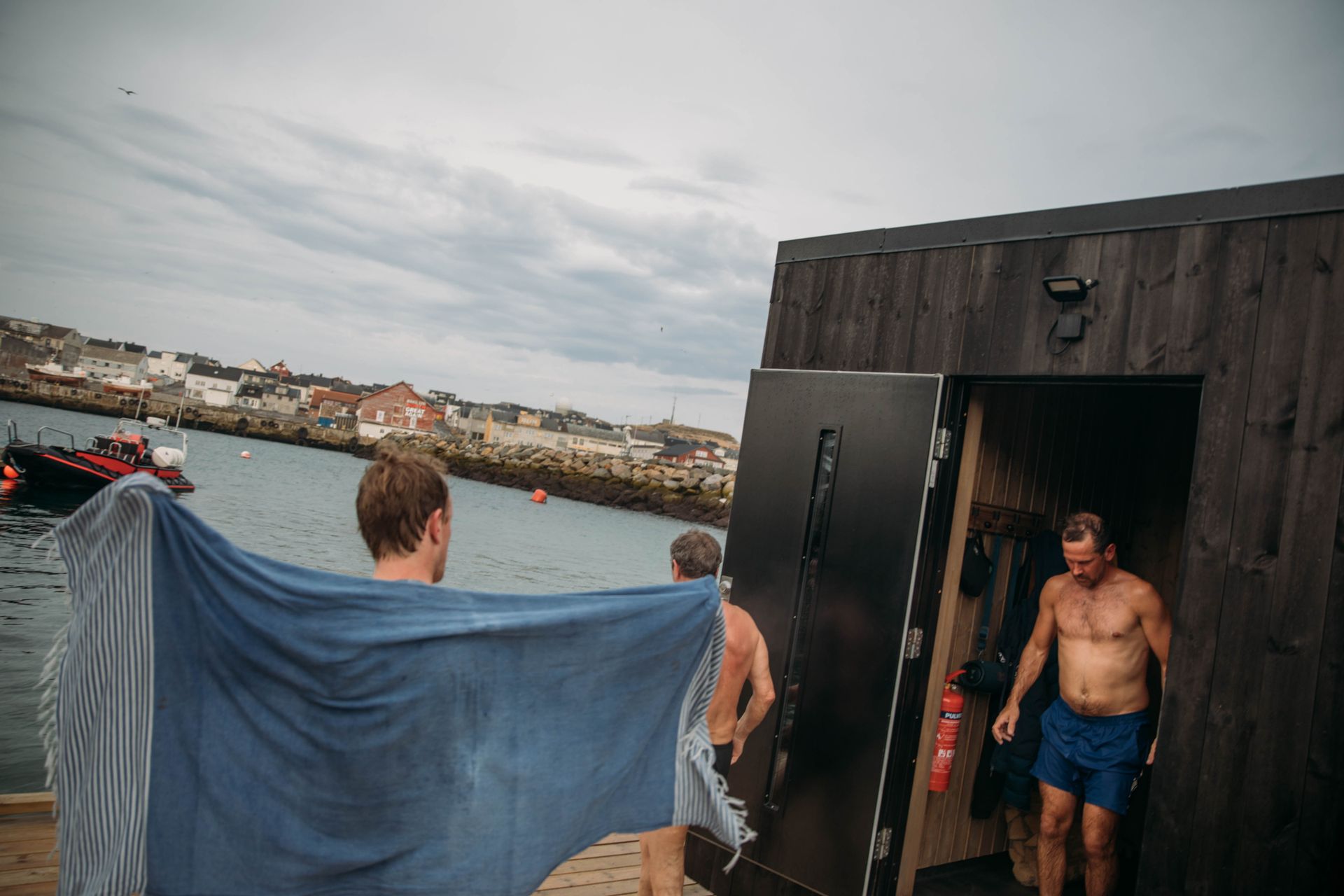 Flere menn står å kjøler seg ned utenfor saunaen på en saunaflåte, man kan se havet.