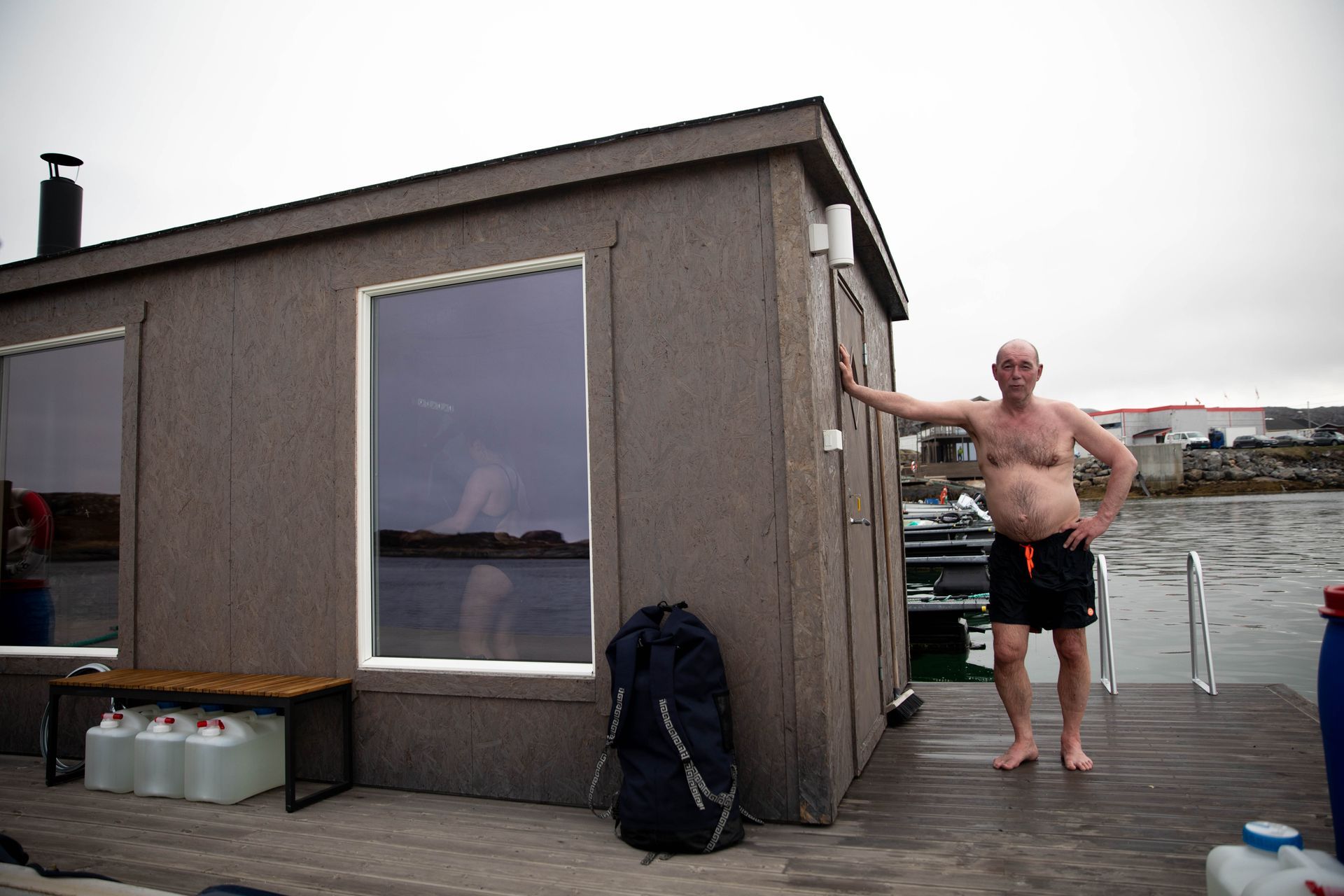 En mann i badebukse lener seg inntil en flytesauna, det står en kvinne ved vinduet i omkledningsrommet.