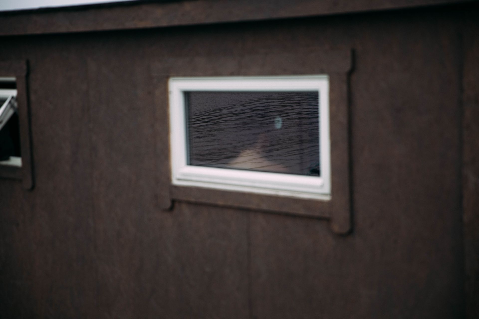 En saunaflåte fotografert utenfra, man kan se ryggen til en mann inne i saunaen gjennom vinduet, havet speiler seg i vinduet.