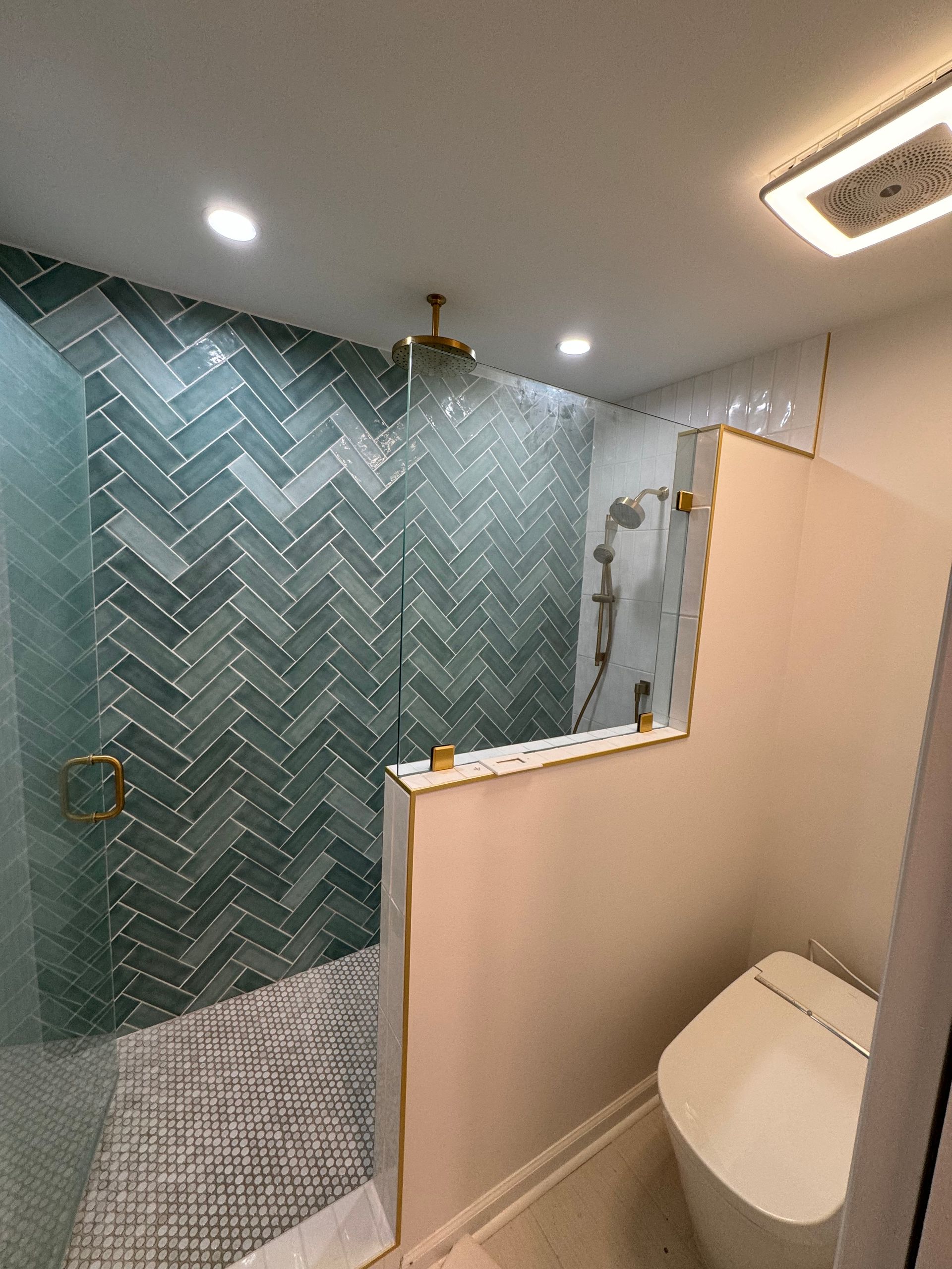 Small bathroom with teal tile shower, beige partition, and toilet under recessed ceiling lights.