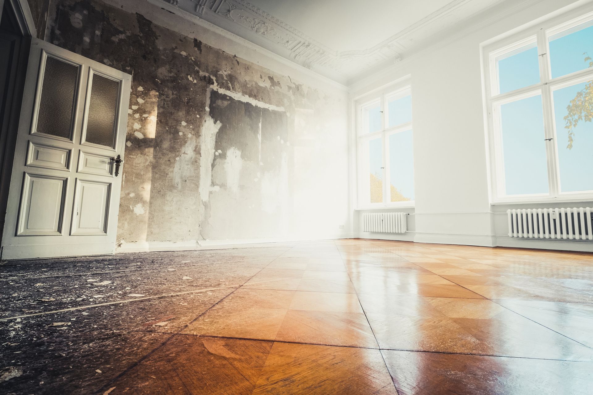 Room undergoing renovation with exposed wall, bright sunlight, and wooden floor.