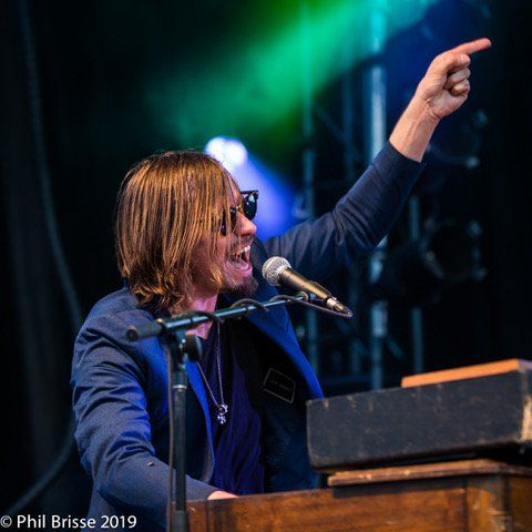 Lacey Doley sitting at keyboard on stage pointing his index while speaking or singing in the mic